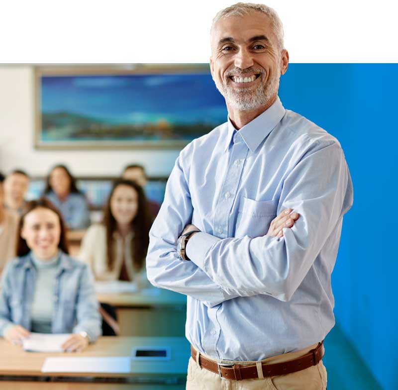 A smiling instructor stands at the front of a classroom with arms crossed, while students sit at desks in the background, slightly out of focus.