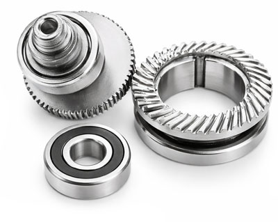 Close-up of precision-machined metal components, including a cylindrical gear assembly, a bearing, and a ring with angled teeth, all with a polished steel finish on a light background.