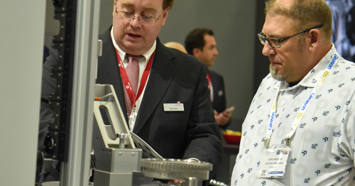 Two men wearing conference badges examine an industrial machine at a trade show booth, with one gesturing toward a component while explaining it.