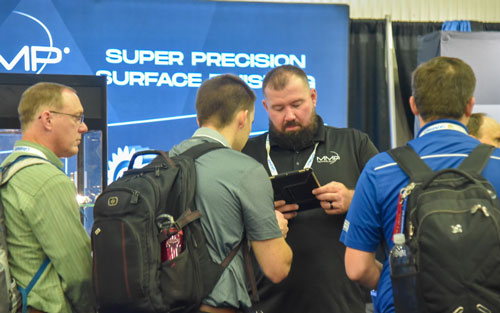 Group of four men gathered at a trade show booth, looking at a handheld device while wearing backpacks and conference badges, with a display behind them promoting super precision surface products.