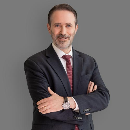 Professional headshot of a man in a dark suit and red tie, standing with arms crossed and smiling against a neutral gray background.