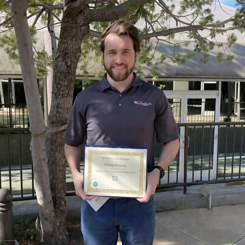 A man stands outdoors holding a framed certificate, smiling in front of a tree and building.