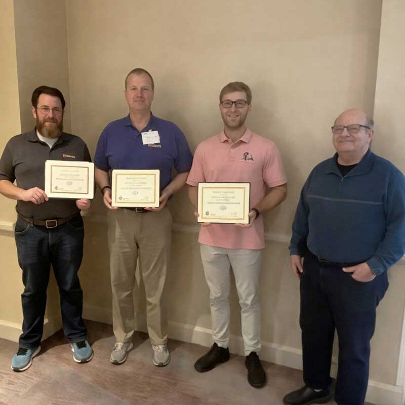 Four men stand indoors against a wall, three holding certificates while posing together for a group photo.
