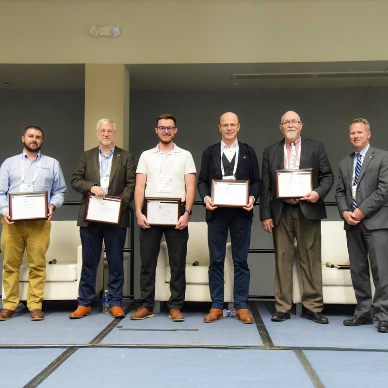 Group of seven men standing on a stage holding framed certificates, posing together during a formal recognition ceremony.