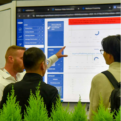 Three people standing in front of a large digital dashboard display, with one person pointing at data visualizations and charts on the screen while the others observe.