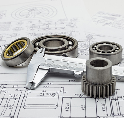 Mechanical components including bearings and a small gear placed on top of technical engineering drawings, with a caliper measuring tool in the foreground.