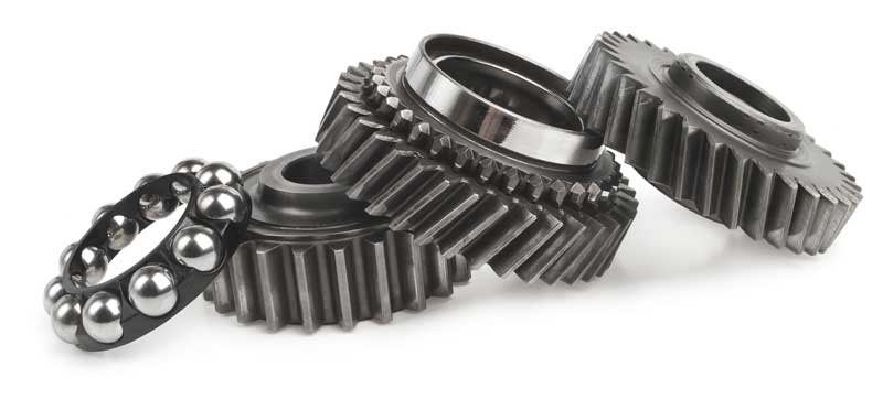 Close-up of several metal gears and a ball bearing, including spur gears and loose bearing balls, arranged together with a polished steel finish on a light background.